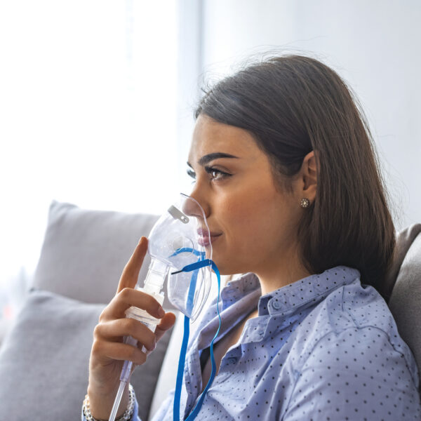 Use nebulizer and inhaler for the treatment. Young woman inhaling through inhaler mask lying on the couch. Woman using asthma machine at home